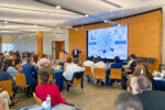 A presenter in a conference room is addressing an audience. The presentation slide features topics relating to "One Health," including environmental, physical, and social elements, with a focus on brain capital.