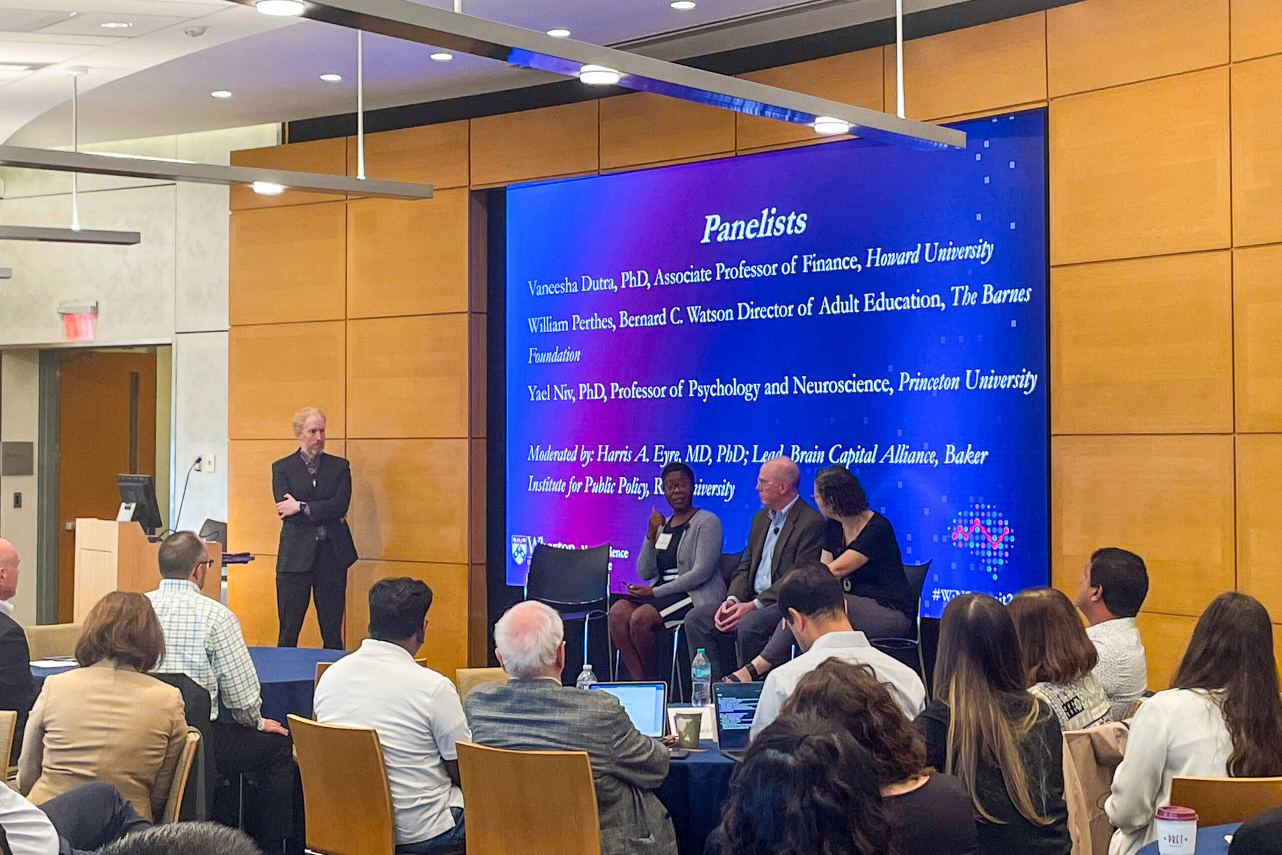 A panel discussion in progress. Several panelists are seated on stage with a moderator standing nearby. A large screen displays information about the panelists and moderator. Audience members are seated, listening attentively.