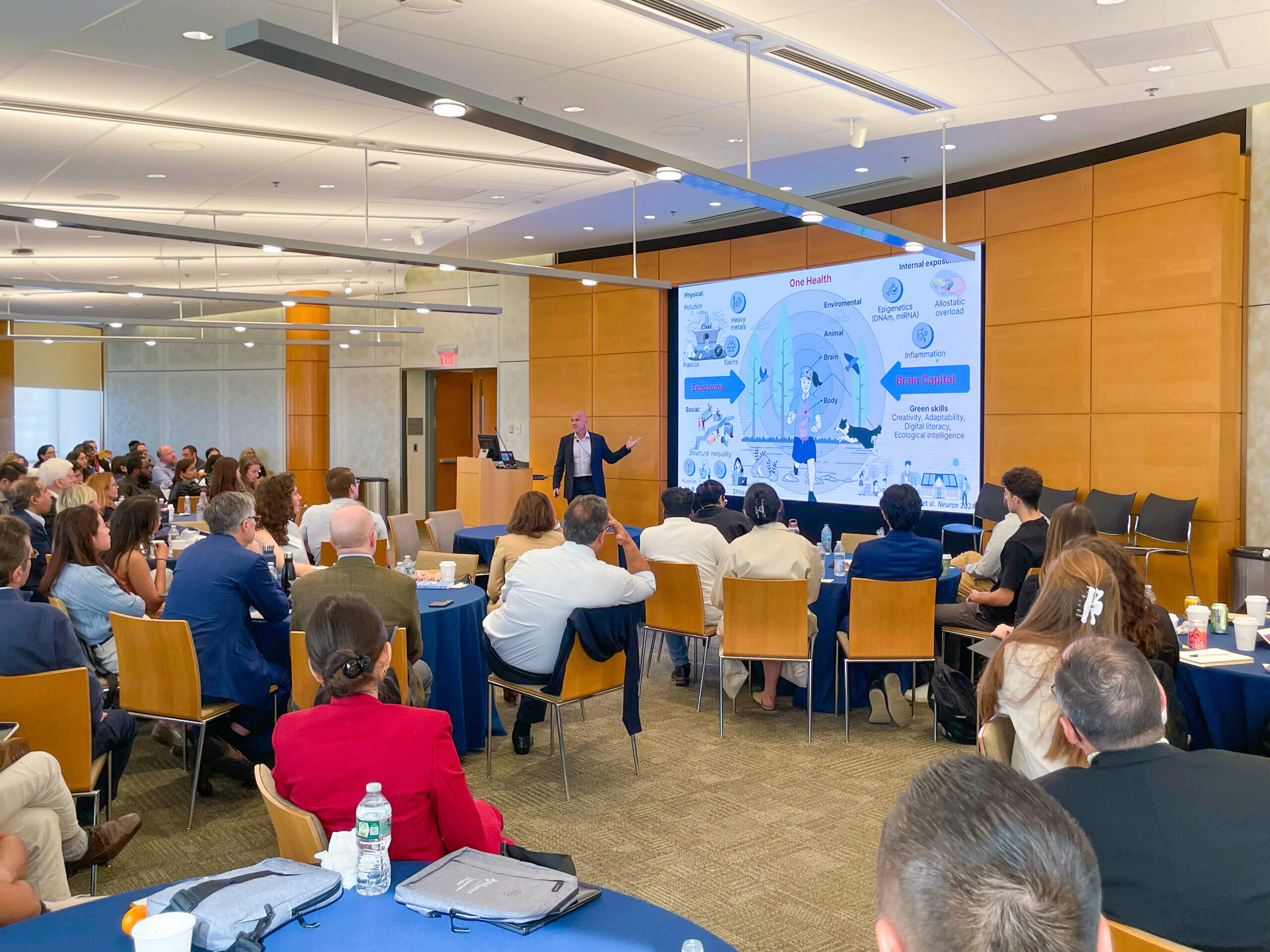 A presenter in a conference room is addressing an audience. The presentation slide features topics relating to "One Health," including environmental, physical, and social elements, with a focus on brain capital.