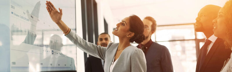 A group of professionals analyzing data on a screen, with one person pointing to a chart. It represents teamwork and data analysis.